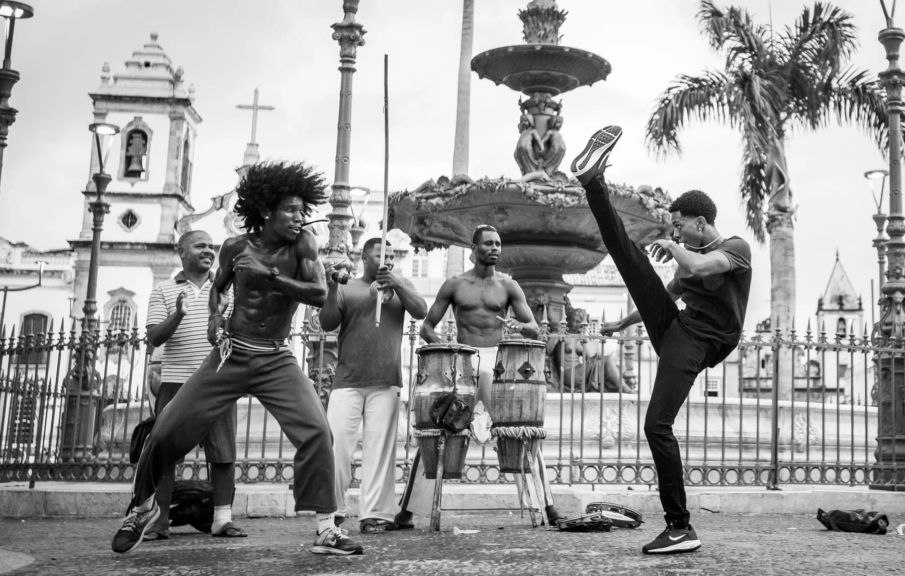 Dois capoeiristas em jogo na rua, acompanhados por berimbau e atabaques